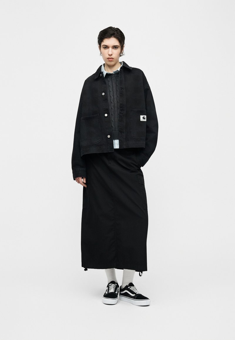 Young person standing wearing a black overshirt, dark cable-knit sweater, long black skirt, white socks, and black-and-white sneakers.