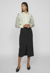 Kraagshirt in crème met zwart bloemenmouwen, gecombineerd met een zwarte rok tot de knie met een split aan de voorkant, afgewerkt met zwarte hakken.