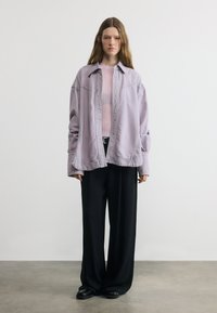 Νεαρή γυναίκα με μακριά καστανά μαλλιά φοράει φαρδιά λεβαντόχρωμη ζακέτα, ανοιχτό ροζ ριμπ πουκάμισο, μαύρο φαρδύ παντελόνι και μαύρα παπούτσια, στέκεται σε εσωτερικό χώρο.