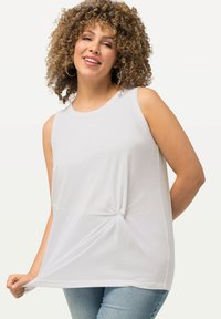 Witte mouwloze top van zachte stof met een geknoopt detail bij de taille, ronde halslijn en een relaxte pasvorm, gecombineerd met lichtblauwe jeans.