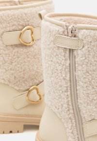 Beige boots with a fluffy fabric upper, gold heart-shaped hardware accents, and side zippers. Textured and smooth surfaces combine.