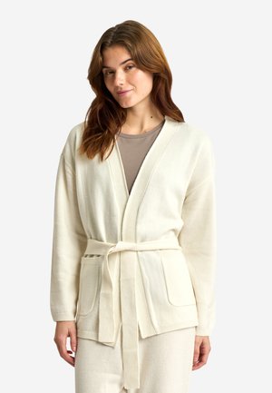 Kvinde med lyst brunt hår iført en cremefarvet cardigan med bælte over en beige top, stående mod en ensfarvet hvid baggrund.