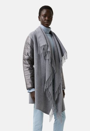 Grå tørklæde lavet af blødt stof, med frynser i kanten. Båret over en grå polstret jakke, med en lyseblå turtleneck under.