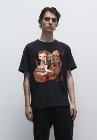 Černé tričko s detailním grafikou postavy a draka v červených a oranžových odstínech. Krátké rukávy a volný střih.