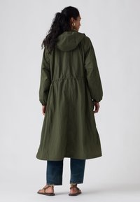 Longue veste vert olive avec capuche, taille ajustable et poignets élastiques, fabriquée en tissu léger, portée par-dessus un jean en denim et des sandales.
