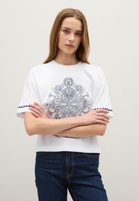 Witte t-shirt met korte mouwen, versierd met gedetailleerde blauwe bloemenborduursels en marineblauwe afwerking op de mouwen, gecombineerd met donkere spijkerbroek.
