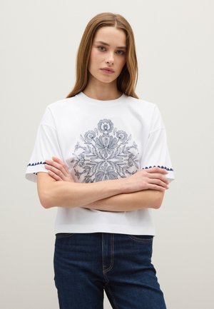 Witte t-shirt met korte mouwen, versierd met gedetailleerde blauwe bloemenborduursels en marineblauwe afwerking op de mouwen, gecombineerd met donkere spijkerbroek.