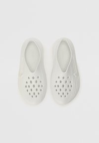 Scarpe bianche slip-on con design perforato, forma arrotondata e liscia, lati texturizzati e un logo discreto sul davanti.