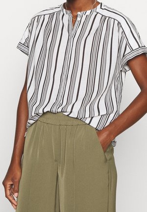 Volwassen persoon draagt een wit shirt met korte mouwen en zwarte verticale strepen, ingestopt in olijfgroene broek met elastische taille, met één hand in de zak.