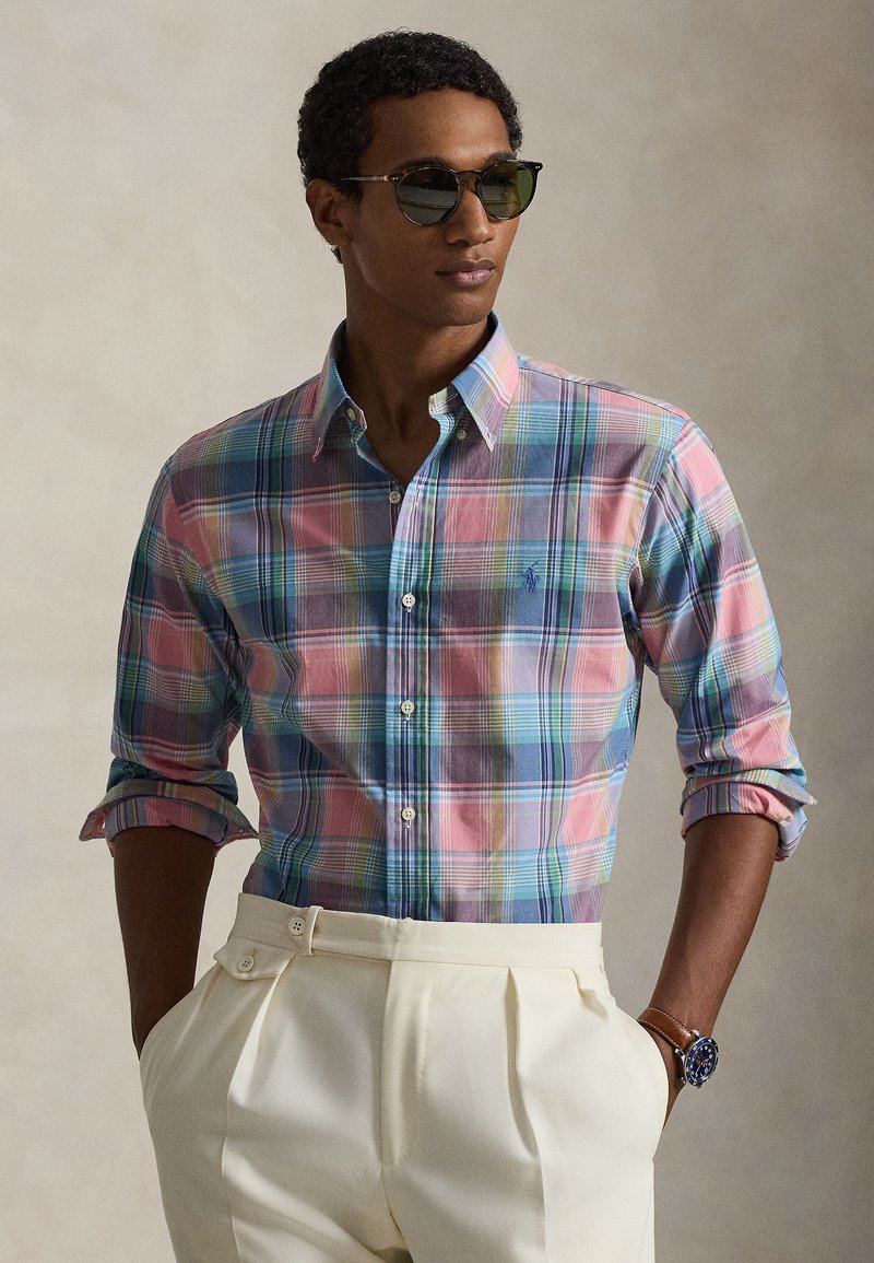 Multicolor plaid shirt featuring light pink, blue, and purple hues, with a button-down collar and rolled sleeves. Worn with white pleated trousers.