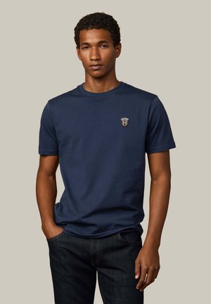 Camiseta de algodón azul marino con mangas cortas, escote redondo y un logotipo de emblema en el lado izquierdo del pecho. Corte ajustado con una textura suave.