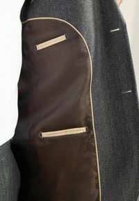 Innenfutter einer grauen Fischgrätenjacke, mit einem glatten braunen Futter, zwei horizontalen Taschen und beigen Akzenten. Dezente Nähte sind sichtbar.