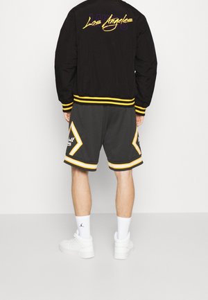 Osoba ubrana w czarną kurtkę Los Angeles z żółtymi akcentami oraz pasujące czarno-żółte szorty, białe skarpetki i białe trampki, stojąca tyłem.