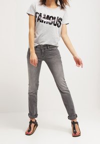 T-shirt grigia con "FAMOUS" in paillettes nere, abbinata a jeans grigi e infradito neri. I jeans sono slim-fit con risvolti.