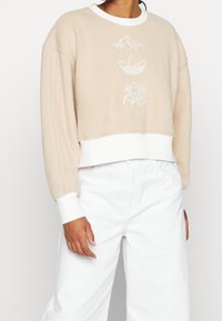 Beige kortad sweatshirt i mjukt tyg med vita ribbade manschetter, prydd med broderade berg, logotyp och blommönster på framsidan.