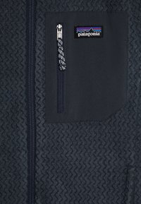 Veste en polaire gris foncé avec un motif texturé, une poche zippée et un patch logo Patagonia. Le zip est doté d'un tirette décorée.