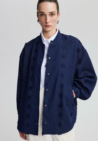 Navy blue textured jacket with floral accents, snap buttons, and relaxed fit. Features drop shoulders and side pockets. White shirt underneath.