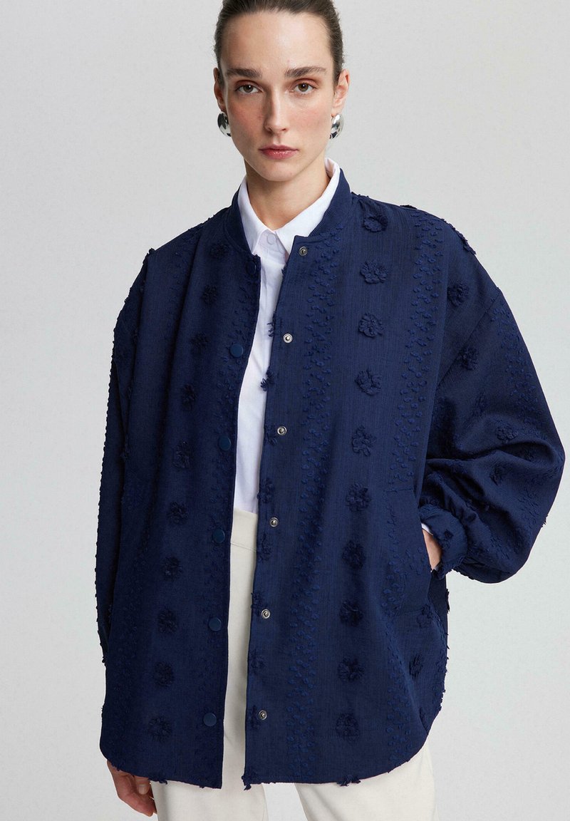 Navy blue textured jacket with floral accents, snap buttons, and relaxed fit. Features drop shoulders and side pockets. White shirt underneath.