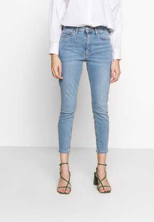 Personne portant un jean skinny bleu clair, une chemise blanche à manches longues et des sandales vertes à talons hauts avec des brides, debout devant un fond uni.