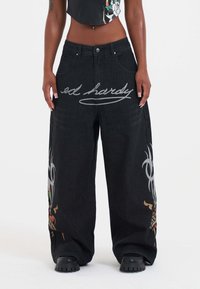 Baggy svarta denimjeans med grafiska mönster som har färgglada motiv och "ed hardy" i kursiv stil på framsidan. Strukturerad tyg, vid passform.