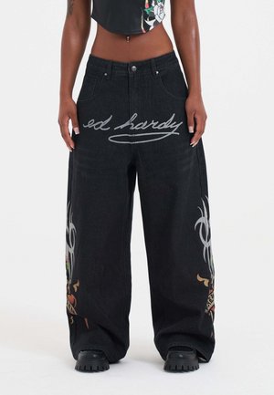 Baggy black denim jeans with graphic designs featuring colorful motifs and "ed hardy" in cursive on the front. Textured fabric, wide-legged fit.