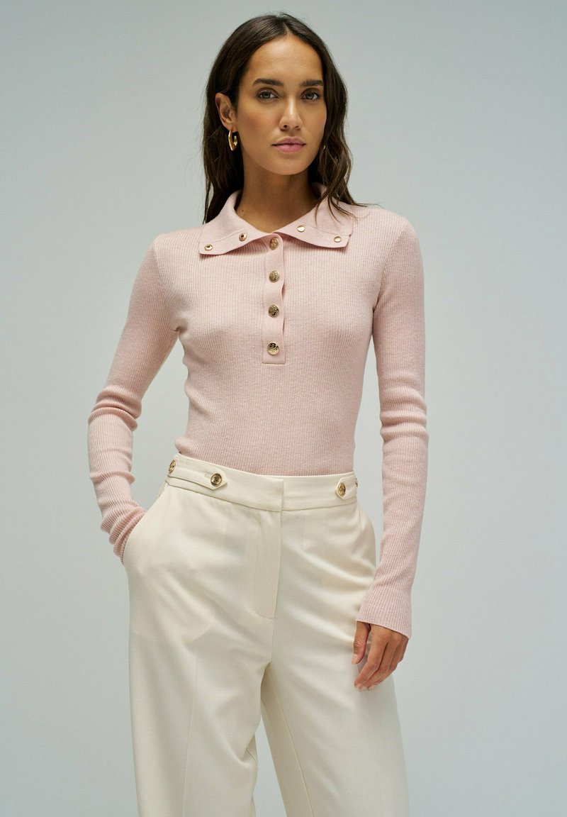 Vrouw met lang donker haar, gekleed in een lichtroze geribbelde top met lange mouwen en gouden knopen, en een hooggesneden crème broek met gouden knoopdetails.