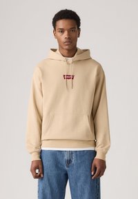 Beige hoodie made of cotton with a front pocket and drawstring hood. Features a red logo patch on the chest and ribbed cuffs. Paired with blue jeans.