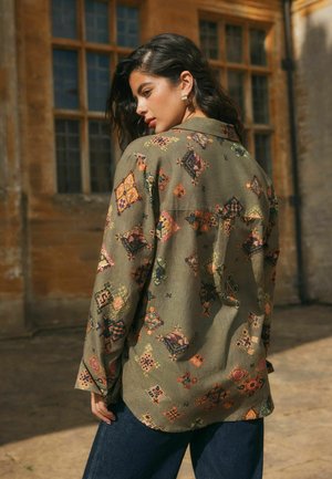 Frau mit dunklem, welligem Haar trägt ein olivgrünes Hemd mit bunten geometrischen Mustern, steht im Freien in der Nähe eines Gebäudes mit großen Fenstern.