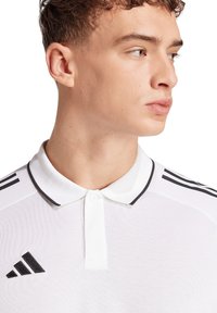 Polo bianco con un motivo testurizzato, rifinitura nera del colletto, caratterizzato da tre strisce nere sulla spalla e un logo Adidas sul petto.