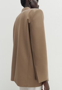 Person iført en beige blazer og hvide bukser, vist fra siden med en neutral baggrund og synlig guldørering.
