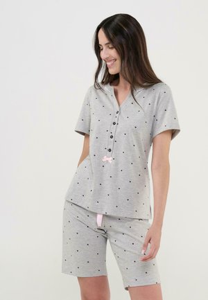 Mujer sonriendo y mirando hacia abajo, vistiendo un pijama gris de manga corta con botones y pantalones cortos a juego con pequeños estampados de corazones negros.