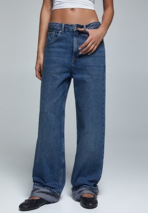 Gezicht vanaf de taille naar beneden van een persoon die losse, wijde blauwe jeans draagt met opgerolde pijpen, zwarte balletschoenen en een wit cropped topje.