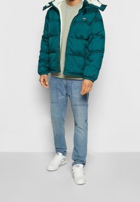 Tyrkysová péřová bunda s bílým fleecem uvnitř, zapínání na zip, elastické manžety, nošená přes světle zelenou košili a modré džíny, doplněná o bílé tenisky.