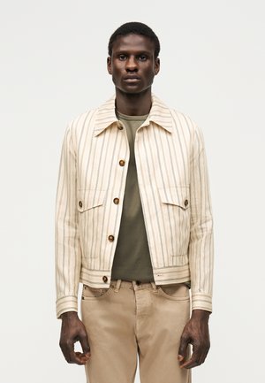 Mand iført en beige vertikalt striped jakke over en olivengrøn skjorte og beige bukser, stående mod en ensartet lys baggrund.