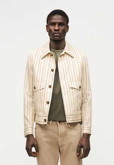 Homme portant une veste beige à rayures verticales sur une chemise vert olive et un pantalon beige, debout devant un fond clair uni.