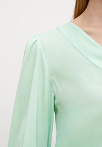 Blusa verde menta con scollatura a V e maniche a sbuffo arricciate. Tessuto liscio e trasparente con una morbida texture e un design fluido.