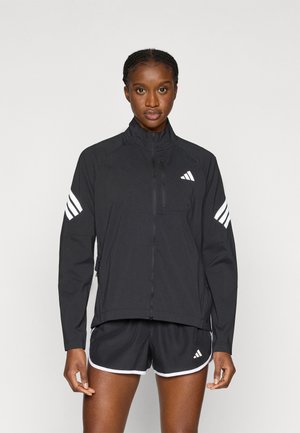 Chaqueta deportiva negra con cuello alto, cremallera frontal y rayas blancas en las mangas. Combinada con pantalones cortos negros con ribete blanco.