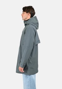Veste imperméable grise avec capuche, présentant une coupe décontractée, de grandes poches latérales et une texture lisse. Portée sur un jean bleu clair.