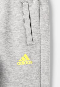 Harmaat collegeshousut pehmeällä pinnalla, sivutaskulla ja kirkkaalla keltaisella Adidas-logolla vasemmassa alareunassa.