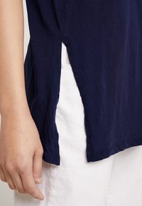 Námorně modrý bavlněný top s bočními rozparky, uvolněný střih, krátké rukávy a kulatý výstřih. Nosený přes bílé kalhoty, zvýrazňující texturu látky.