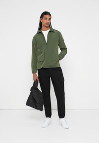 Olijfgroene lichtgewicht jas met een rits aan de voorkant, twee voorzakken en een zwart accentlabel, gedragen over een wit t-shirt en zwarte broek.
