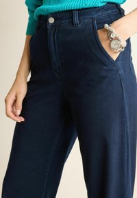 Pantalons taille haute bleu foncé en denim, avec des poches avant et un accent en bouton argenté. Texture lisse, design à jambe droite.
