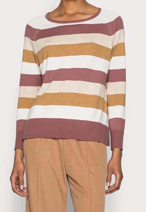 Personne portant un pull à manches longues avec de larges rayures horizontales beige, marron, blanc et rose pâle, assorti à un pantalon marron clair.