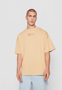 Beige oversized bomull t-skjorte med korte ermer og rund hals, med en subtil brodert logo i brun på brystet.