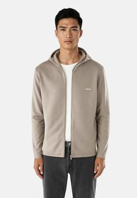 Lys grå zip-up hettegenser med glatt tekstur, snorhette og hvit logo på venstre bryst, kombinert med mørke jeans.