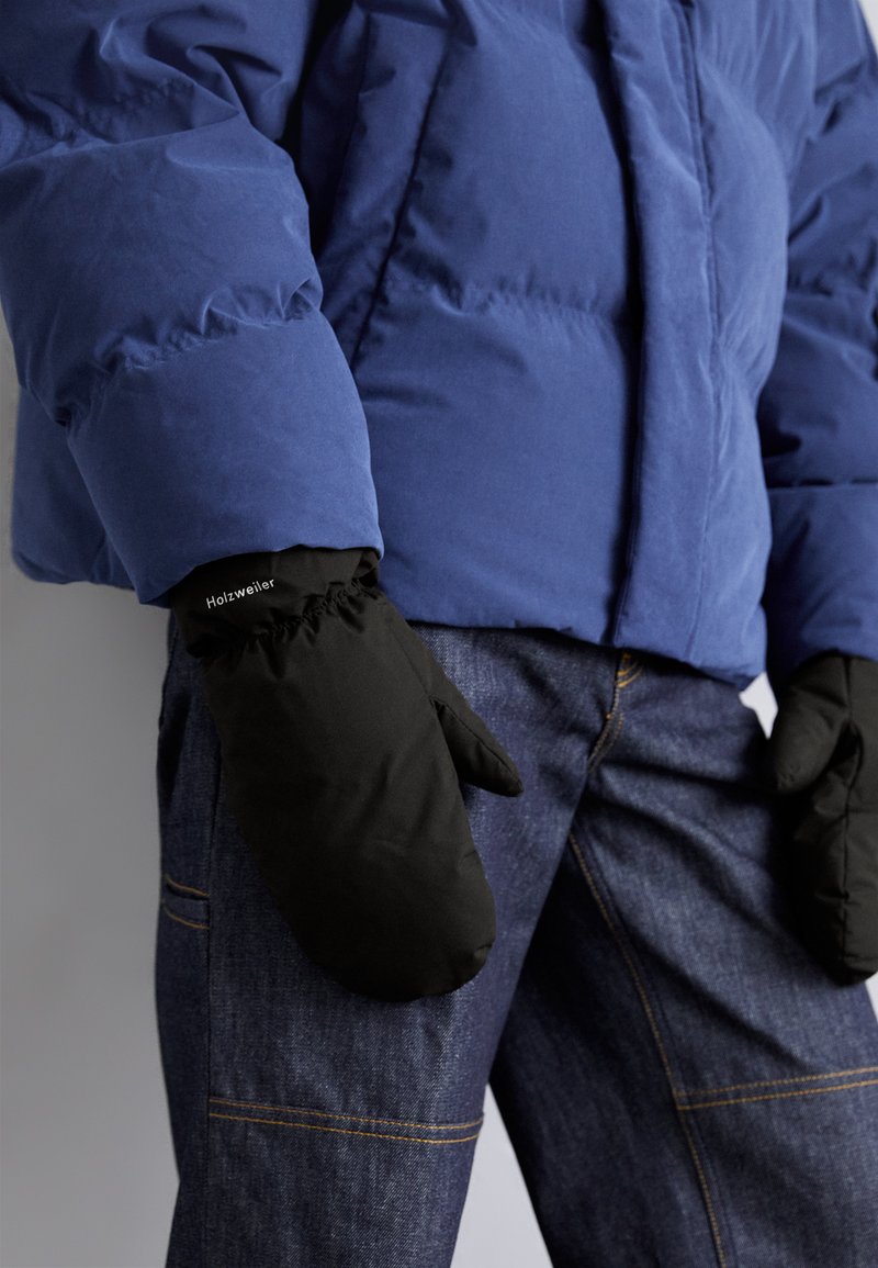Person trägt eine blaue Steppjacke, schwarze isolierte Fäustlinge mit der Aufschrift "Holzweiler" und dunkelblaue Jeans mit sichtbaren Nähten.