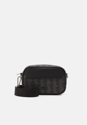 Petit sac bandoulière noir avec motif géométrique, fermeture éclair sur le dessus, sangle large amovible et nom de marque discret sur le panneau avant.