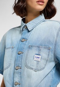 Ljusblå denimskjorta med krage, sex framknappar med "Lee"-logotyp och två bröstfickor med en "Lee Workwear"-etikett.