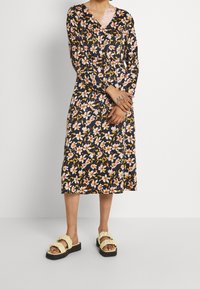 Květinové šaty s dlouhými rukávy, véčkovým výstřihem, námořnickým pozadím a oranžovými květinami. Dokončeno s žlutými sandály s volány a tmavou platformou.