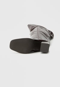 Brązowy, sięgający kolan, skórzany but z obcasem typu słupek, miękką fakturą i gładkim wykończeniem. Podeszwa jest antypoślizgowa, z teksturowanym wzorem.